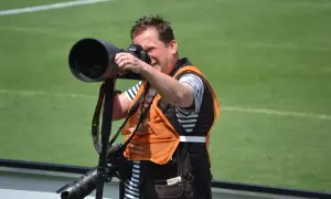 Fotograf z aparatem na monopody, w pomarańczowej kamizelce, gotowy do uchwycenia akcji na boisku.