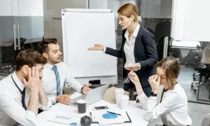 En kvinna gestikulerar mot en whiteboard medan kollegor ser utmattade ut. Känslan av manipulativt beteende hänger i luften under mötet.