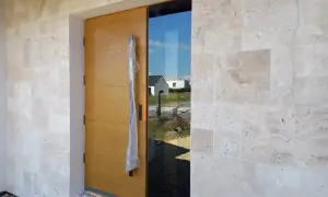 Eleganckie drzwi z drewna i szkła, otoczone ścianą z jasnego trawertynu, zapraszają do nowoczesnego domu.