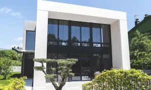 Moderna casa con fachada gris y beige, grandes ventanales y jardín cuidado.