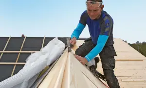 A roofer wearing safety glasses hammers a wooden plank onto a roof under construction.