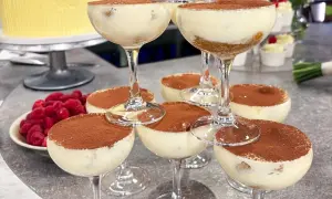 Flera glas med krämig tiramisu, pudrad med kakao. Hallon och en gul tårta i bakgrunden.
