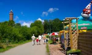Rodzina spaceruje ścieżką w Ustroniu Morskim w kierunku latarni. W tle wesoły klaun i zielone parasole.