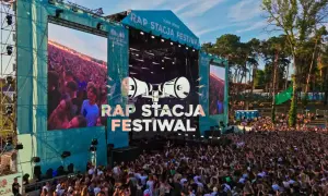 Tłum ludzi na koncercie rap festiwal. Na scenie wielkie ekrany z napisem "RAP STACJA FESTIWAL" i grafiką megafonów.
