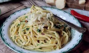 En tallrik med spaghetti aglio e olio, toppad med riven parmesan och persilja. En röd chili och ostbitar syns i bakgrunden.