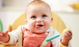 Uśmiechnięte dziecko w krzesełku, z językiem na wierzchu, trzymające łyżeczkę. Zastanawiasz się, ile powinno jeść 5-miesięczne dziecko?