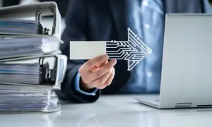Ręka trzyma kartkę z cyfrową strzałką, obok sterty dokumentów i laptopa. Nowoczesny dowód księgowy.