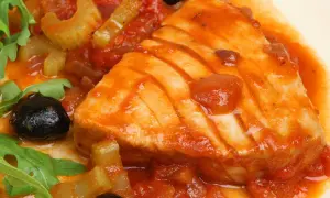 Un succulento tonno in padella con pomodoro, olive nere e sedano, guarnito con rucola fresca.