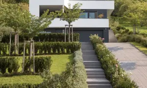 Moderner Garten am Hang mit gepflasterter Auffahrt, Treppen und gepflegten Hecken. Ein Haus mit weißen Fassaden und dunklen Fenstern dominiert den Hintergrund.