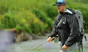 Ein Mann mit Hut und Sonnenbrille steht im Wasser und übt das **streamer fischen**. Er trägt eine schwarze Regenjacke und einen Rucksack.