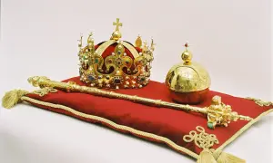 Złote klejnoty koronne: korona, berło i jabłko królewskie, ozdobione drogimi kamieniami i perłami, spoczywają na czerwonej poduszce.
