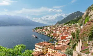 Vista panoramica di una città con tetti rossi e case colorate, incastonata tra la montagna e il blu del lago di Garda. Una nave solca le acque tranquille.