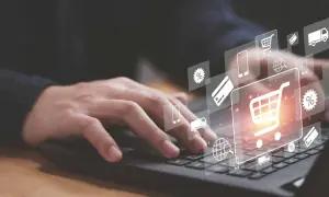 Dłoń na klawiaturze laptopa, nad nią ikony symbolizujące e-commerce: koszyk, karta płatnicza, ciężarówka, procenty, globus.