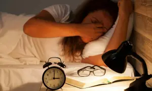 En person som gnuggar sig i ögonen i sängen, med en väckarklocka, glasögon och en bok på nattduksbordet. Känns som att vaknar flera gånger per natt.
