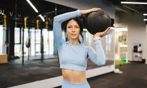 Kobieta w niebieskim stroju sportowym trzyma czarną piłkę nad głową. To ćwiczenie, które może być częścią treningu hyrox.