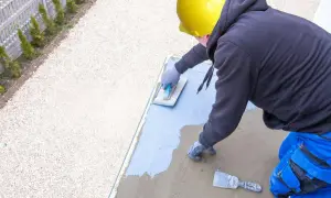 Pracownik w żółtym kasku wykonuje hydroizolację tarasu, nakładając masę izolacyjną na betonową płytę.
