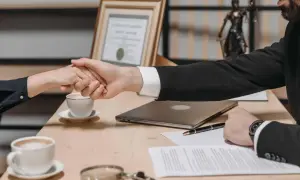Uścisk dłoni między dwoma osobami przy biurku z dokumentami, kawą i laptopem.