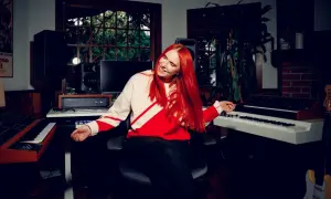 Noonie Bao, with vibrant red hair, smiles and gestures in a music studio filled with synthesizers and speakers.