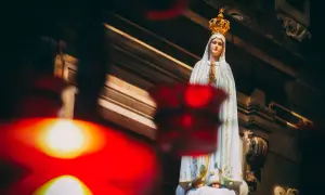 Figurka Matki Bożej Fatimskiej w koronie, z rękami złożonymi do modlitwy. W tle widać architekturę kościoła, a na pierwszym planie rozmyte czerwone światła, symbolizujące trzecia tajemnica fatimska.