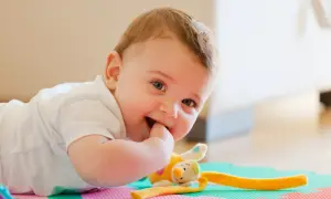 Uśmiechnięte 11-miesięczne dziecko leży na macie, gryząc paluszek i bawiąc się pluszową zabawką.