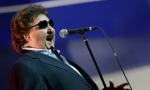 Krzysztof Krawczyk w wieku 60 lat, w okularach przeciwsłonecznych i garniturze, śpiewa do mikrofonu.