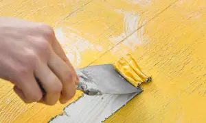 Mano raspando pintura amarilla vieja con una espátula. El decapado de pintura revela madera debajo.