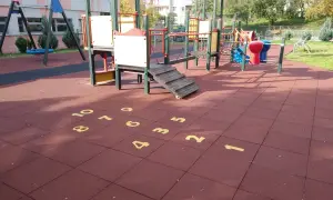 Plac zabaw z huśtawkami i drabinkami. Na czerwonej nawierzchni namalowana jest gra w klasy, a obok niej widać elementy do zabawy w twister.