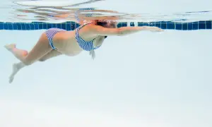 En gravid kvinna i randig bikini simmar framåt i en pool. Vattnet är klart och blått.