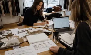 Dos mujeres revisan proyecciones de ventas y calendarios de producción. Una escribe en una libreta, la otra señala un documento. Se preguntan qué significa sin stock.