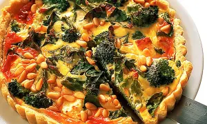 En gyllene vegetarisk paj med broccoli, spenat och pinjenötter. En bit lyfts upp med en pajspade.