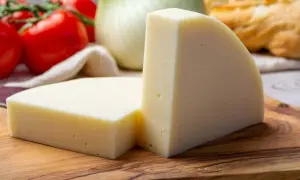 Zwei Stücke heller Käse auf einem Holzbrett, daneben Tomaten und Fenchel.