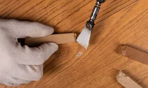 Mano guantata rimuove schegge da un parquet rovinato, mostrando rimedi fai da te per riparare il legno danneggiato.