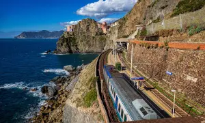 Ein Zug fährt entlang der Küste Italiens am Meer entlang. Bunte Häuser kleben an steilen Felsen.