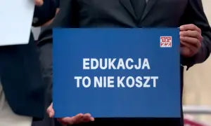 Osoba trzyma niebieską tabliczkę z napisem "EDUKACJA TO NIE KOSZT". Domaga się podwyżki dla nauczycieli 2026.
