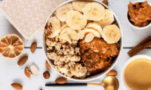 Owsianka z bananem, masłem orzechowym i migdałami w misce, obok suszonej cytryny i kawy.