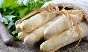Un bouquet d'asperges blanches, prêtes à être poêlées, est noué avec une ficelle sur un tissu. Des herbes fraîches et un couteau sont visibles en arrière-plan.