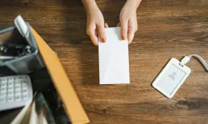 Mani che tengono una busta bianca, forse una raccomandata disdetta affitto, su un tavolo di legno con un badge e una scatola.