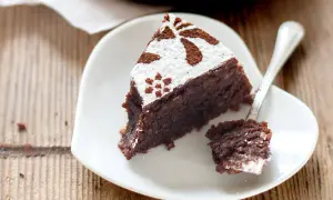 Una fetta di torta caprese ricetta originale napoletana, cosparsa di zucchero a velo e cacao, su un piatto bianco a forma di cuore.