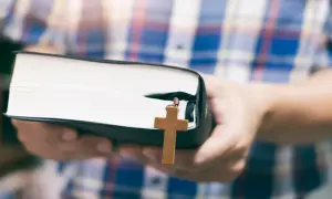Osoba w kratę trzyma Biblię z drewnianym krzyżykiem, symbolizującym dwa przykazania miłości.