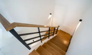 Nowoczesne schody z drewnianymi stopniami i czarną balustradą. Jak oświetlić schody w domu? Dyskretne kinkiety na ścianie tworzą nastrojowy blask.