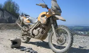 Żółty motocykl po off-roadowej przygodzie, pokryty błotem. Obok leżą kask i rękawice. Idealny do lekkiego enduro.