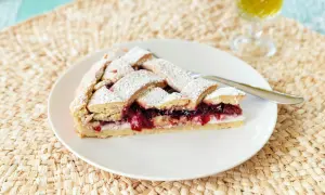 Una fetta di crostata di ricotta con confettura di ciliegie, cosparsa di zucchero a velo, su un piatto bianco.