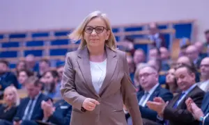 Kobieta w okularach i brązowej marynarce idzie na tle publiczności.