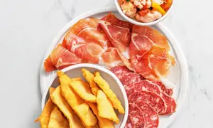 En tallrik med italienska delikatesser: tunt skivad prosciutto, salami och krispiga gnocco fritto. En skål med marinerade grönsaker.