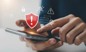 Dłonie trzymające smartfon, nad którym wyświetlają się ikony symbolizujące bezpieczeństwo i ochronę przed zagrożeniami, w tym czerwiona tarcza z wykrzyknikiem. To wizualizacja skutecznego **antywirusa na telefon**.