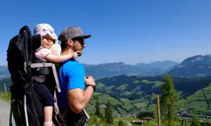 Vater trägt sein Baby in einer Kraxe auf einem Berg. Die Aussicht ist atemberaubend.