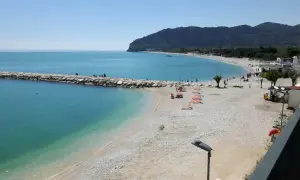 Mattinata serena sulla spiaggia di piana, con il mare azzurro che lambisce la costa ghiaiosa e il molo di sassi.