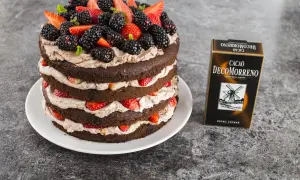 Wielowarstwowy tort czekoladowy z kremem mascarpone, udekorowany świeżymi truskawkami, jeżynami i miętą. Obok stoi opakowanie kakao DecoMorreno.