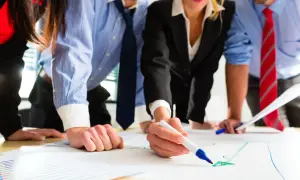 Zespół analizuje wykresy, jeden z pracowników zaznacza coś markerem. Wszyscy skupieni na pracy, tworząc **irr**ytujący projekt.