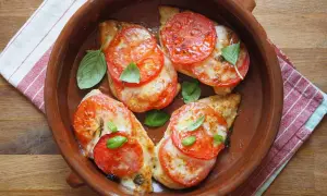 Cztery porcje pieczonej piersi z kurczaka z mozzarellą i pomidorami, udekorowane listkami bazylii, podane w glinianym naczyniu.